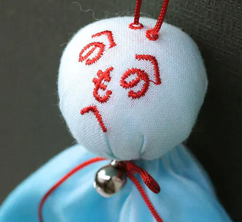 Bambolina teru teru bozu con hiragana rossi sul volto e un campanellino.